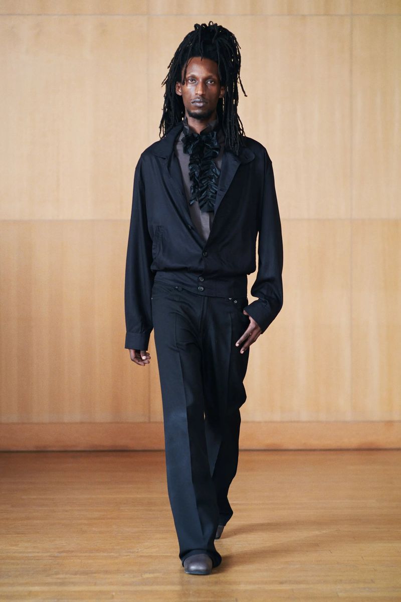Dark-skinned adult male in all-black fashion, walking on a wooden floor in front of a light wood paneled wall, runway style.