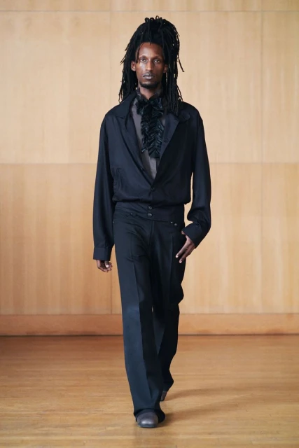 Dark-skinned adult male in all-black fashion, walking on a wooden floor in front of a light wood paneled wall, runway style.