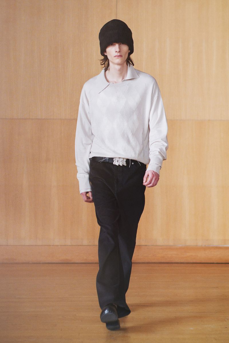A model wearing a light-colored long-sleeved shirt, dark pants, a belt, and a knit cap walks down a light wood runway.