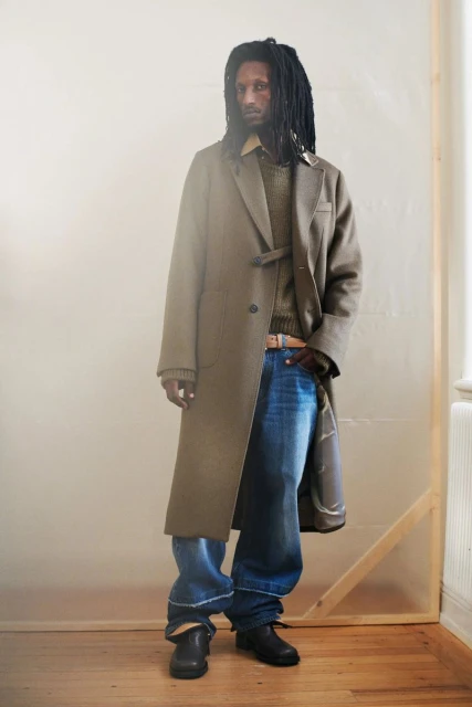 A lone adult male stands in a long olive-green coat and jeans, indoors on a hardwood floor near a window with diffused lighting.