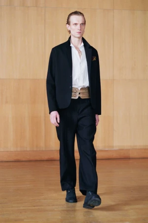 Model walking on a wooden floor in a black blazer, white shirt, and wide trousers, captured for Our Legacy F/W 25.