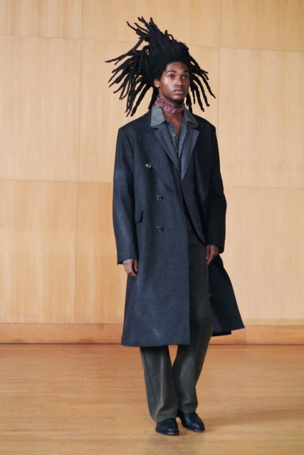 Young adult male model with dreadlocks wearing a double-breasted overcoat, shirt, trousers, and shoes in a studio setting against a wood backdrop.