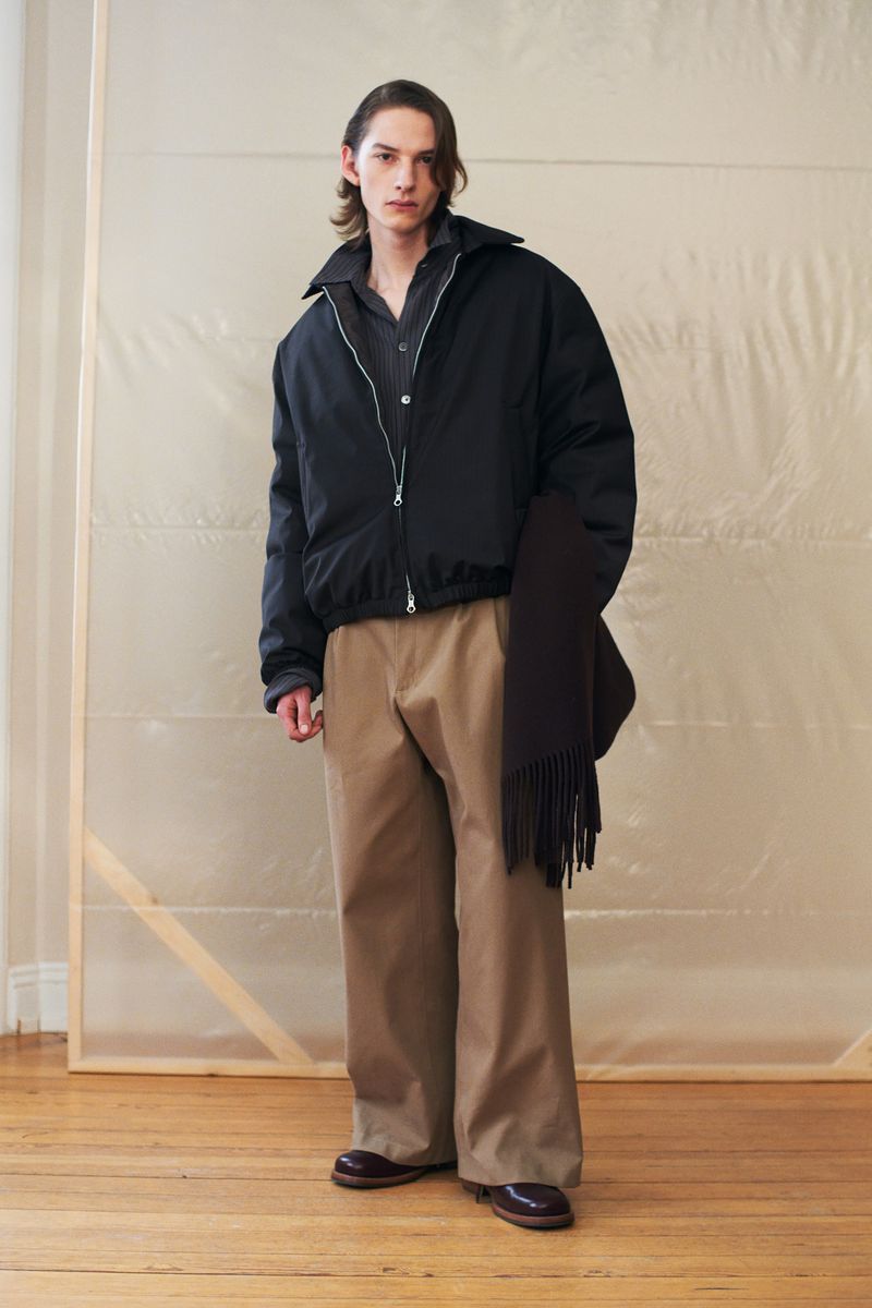 Studio shot of a male model showcasing a black bomber jacket, brown collared shirt, beige trousers, and a dark brown scarf.