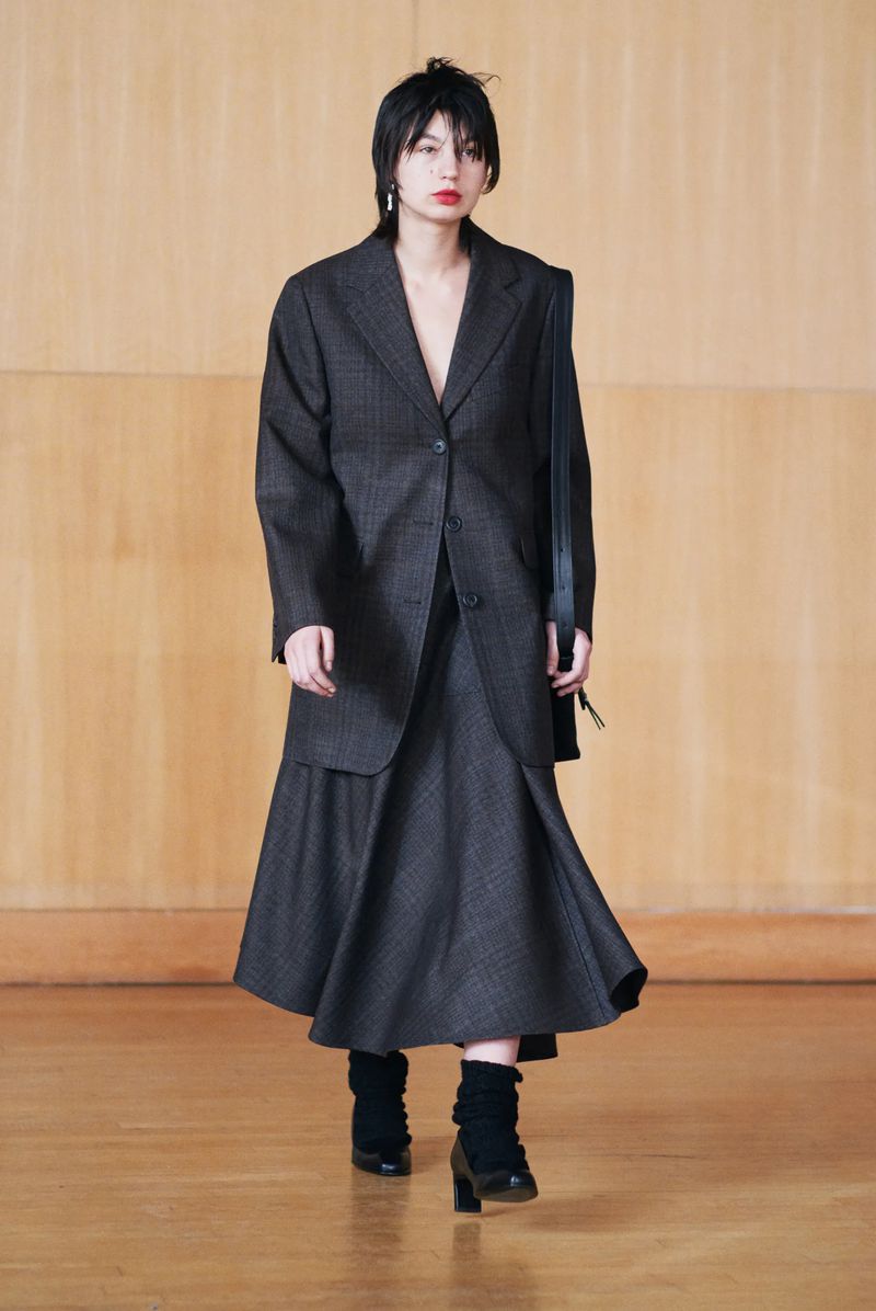 A woman models a blazer and skirt outfit in a neutral tone studio. She is wearing black boots and carrying a handbag on her shoulder.