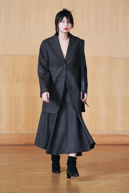 A woman models a blazer and skirt outfit in a neutral tone studio. She is wearing black boots and carrying a handbag on her shoulder.