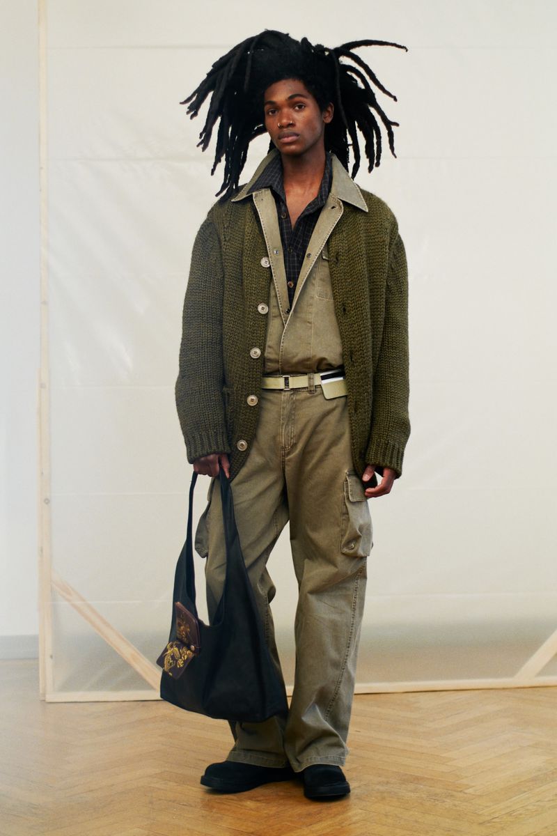Full-length shot of a person in an olive green outfit with an oversized cardigan, standing against a white backdrop with a black tote bag.