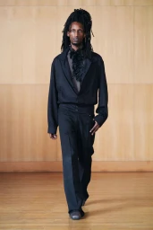 A Black man with long dreadlocks walking in a black jacket and matching trousers against a wooden paneled wall.