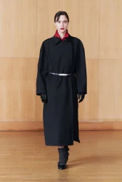 Model in a long black coat with a belt and red collar standing against a wooden panel wall