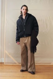 Model wearing a black zip jacket and wide trousers standing in a studio with wooden floors and a translucent backdrop.