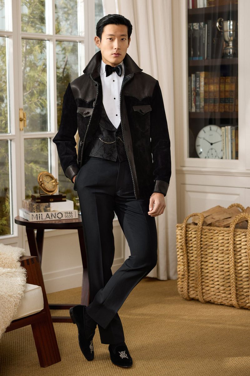Well-dressed Asian male in black jacket, vest, and pants stands indoors near window and bookcase, striking a confident pose.