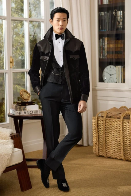 Well-dressed Asian male in black jacket, vest, and pants stands indoors near window and bookcase, striking a confident pose.