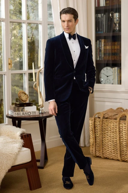 Male fashion model poses indoors wearing a navy blue velvet tuxedo with bow tie, cummerbund, and slip-on shoes in a classic style.