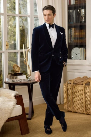Male fashion model poses indoors wearing a navy blue velvet tuxedo with bow tie, cummerbund, and slip-on shoes in a classic style.