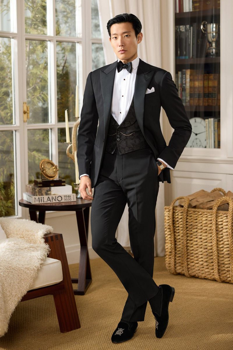 An adult male in a black tuxedo poses in an indoor setting with a classic style. The photo emphasizes formal fashion and elegant presentation.