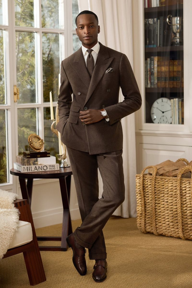 An adult man in a brown suit stands in front of a window and bookcase, looking elegant and composed in an indoor setting.