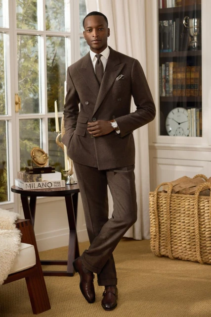 An adult man in a brown suit stands in front of a window and bookcase, looking elegant and composed in an indoor setting.