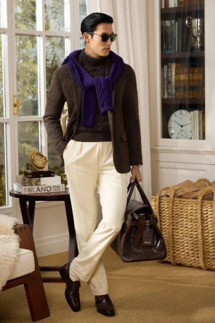 Male fashion model poses indoors wearing a blazer, sweater, scarf, pants, and sunglasses, while holding a leather duffel bag.