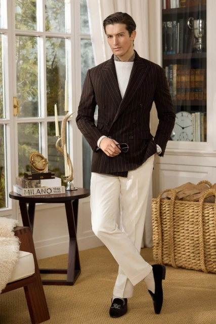 Adult man in a double-breasted pinstripe blazer and cream pants, standing indoors with a warm, inviting ambiance. Fashion editorial.
