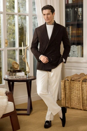 Adult man in a double-breasted pinstripe blazer and cream pants, standing indoors with a warm, inviting ambiance. Fashion editorial.