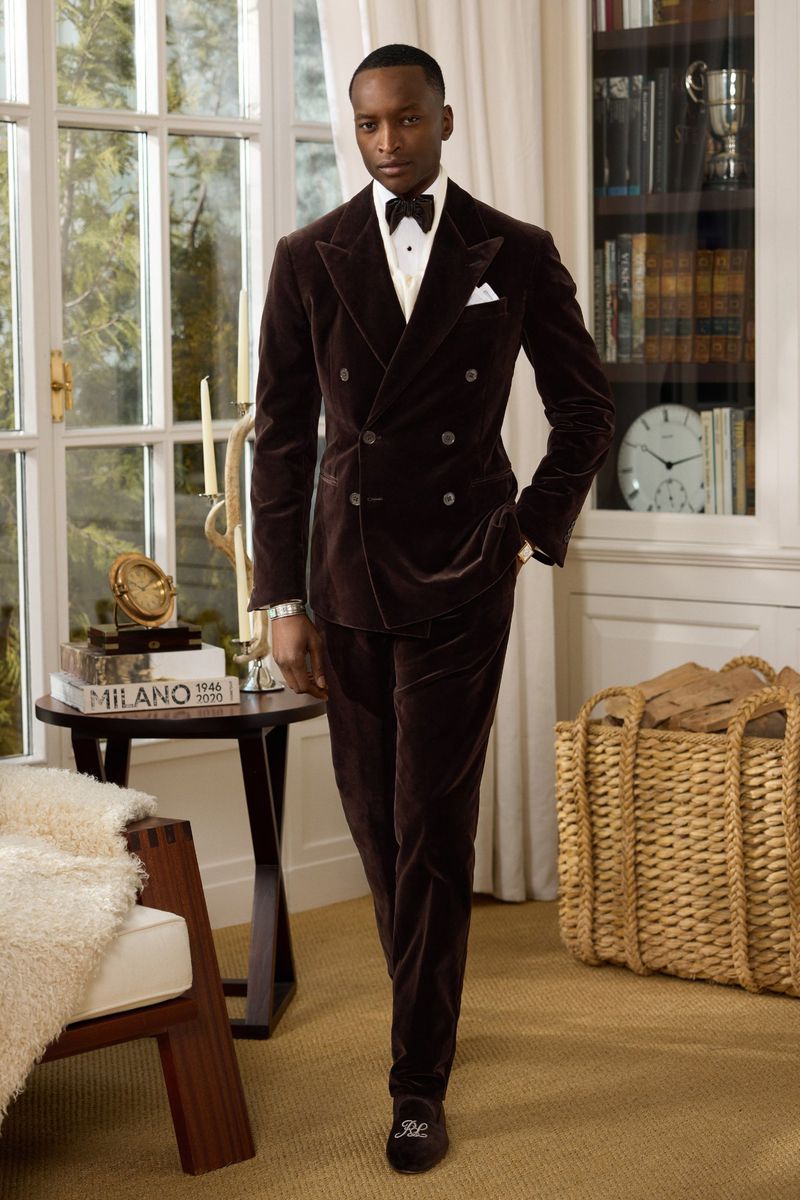 An African American man in a formal velvet suit with a bow tie walks in a stylish interior with a bookcase and natural light.