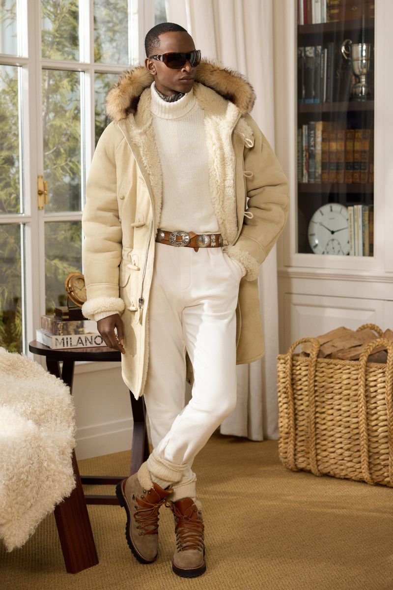 Man in cream turtleneck, white pants, and tan boots with fur-trimmed hooded coat in interior with bookshelf and natural light.