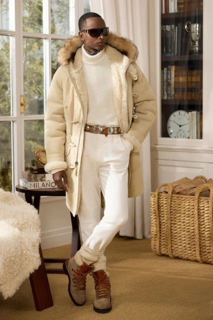 Man in cream turtleneck, white pants, and tan boots with fur-trimmed hooded coat in interior with bookshelf and natural light.