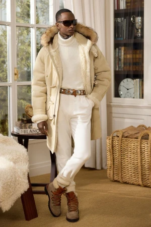 Man in cream turtleneck, white pants, and tan boots with fur-trimmed hooded coat in interior with bookshelf and natural light.