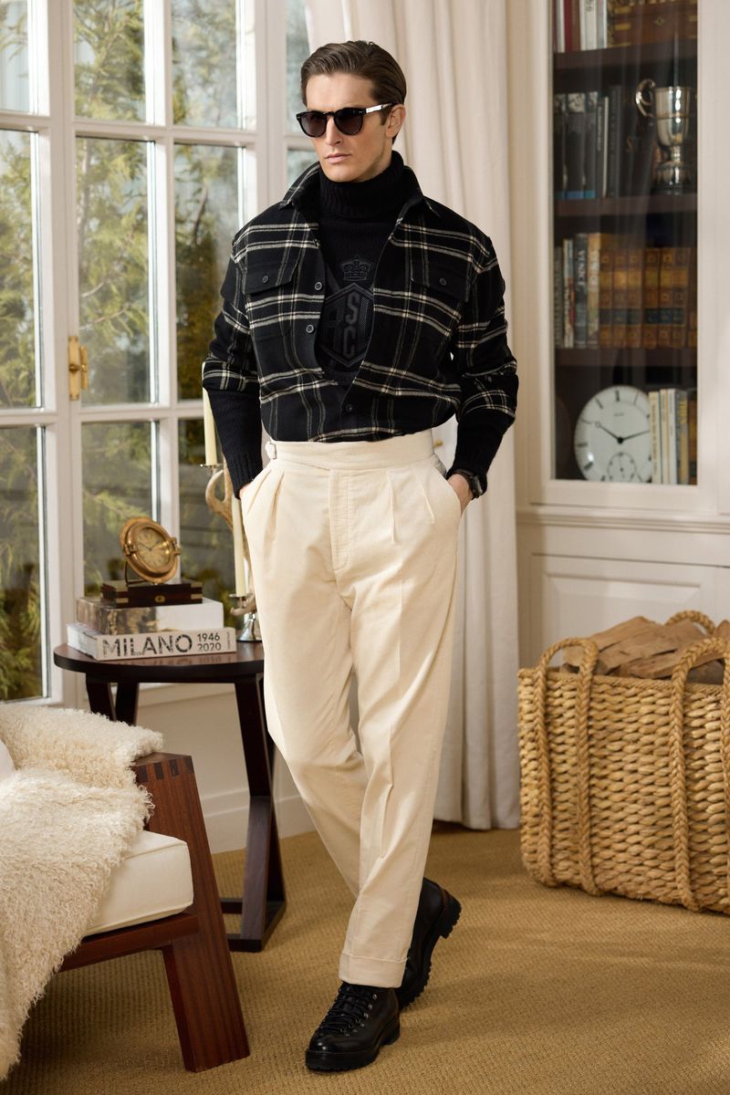 Man wearing a black turtleneck, plaid shirt, and cream trousers posing in an elegant room with a large window and bookshelves.