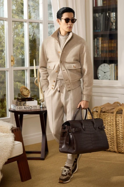 Adult male in a beige jacket and sweatpants carrying a leather duffel bag poses in an indoor setting with warm lighting.