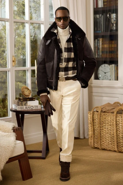A stylish adult male poses indoors wearing a black shearling bomber jacket, a white turtleneck, a plaid shirt, and cream-colored pants.