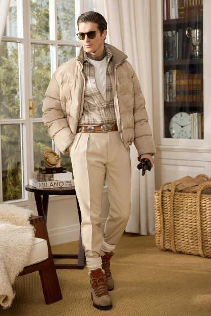 Male fashion model in tan puffer jacket and khaki trousers poses indoors with natural light and classic interior decor.
