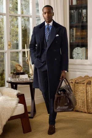 Man wearing a navy coat over a plaid suit and tie holding a leather duffel bag in a luxury interior setting