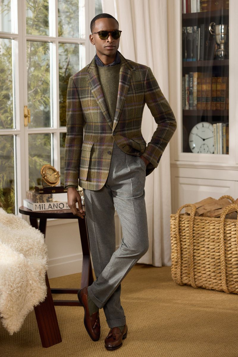Adult male model wearing a brown plaid blazer, olive-green sweater, and gray trousers stands indoors in front of a large window.