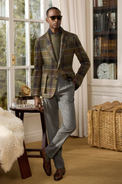 Adult male model wearing a brown plaid blazer, olive-green sweater, and gray trousers stands indoors in front of a large window.