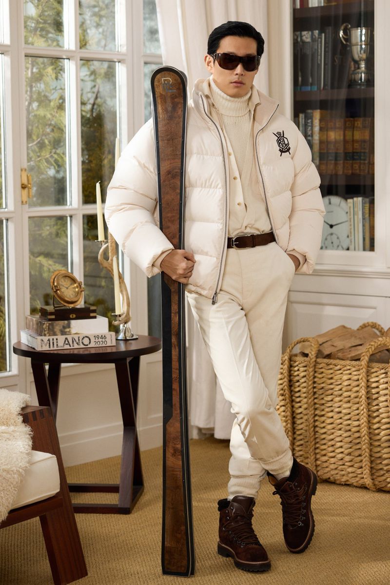 An Asian male poses indoors in a cream puffer jacket, cream pants, and brown boots, holding skis, with books and decor in the background.