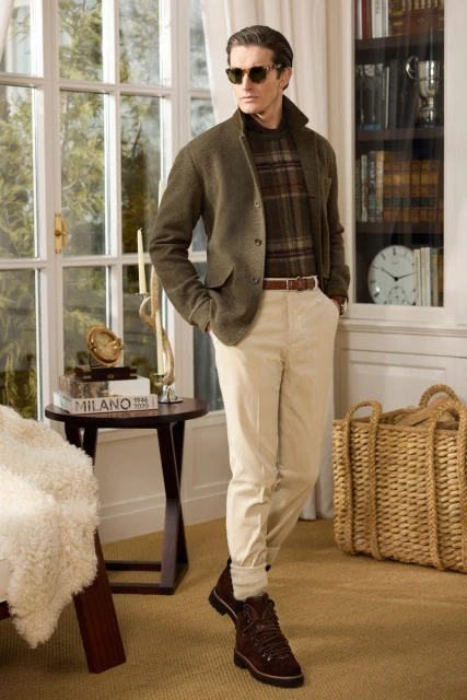 Adult male in an olive blazer, plaid sweater, and khaki pants stands in front of a window, illuminated by natural light in an indoor setting.