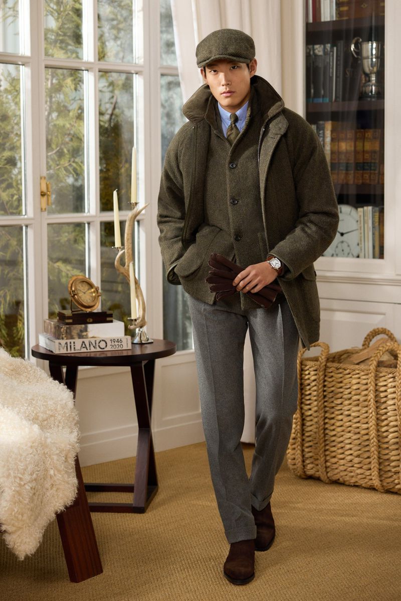Man in tweed outfit walking indoors. He wears a cap, jacket, vest, shirt, and trousers with leather gloves in hand near a side table.