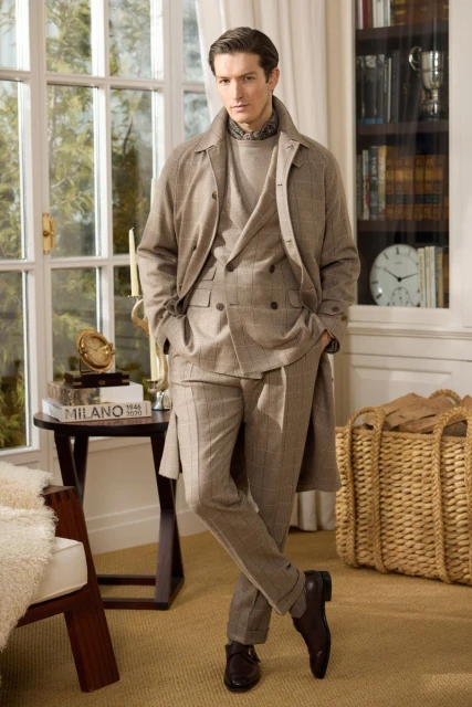 Man in a plaid suit and coat poses indoors near a window and bookcase, showcasing classic menswear fashion in a lifestyle setting.