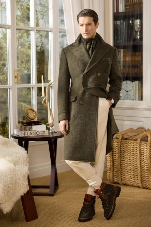 Man wearing a long olive green wool coat, cream trousers, and dark boots standing in a wood-paneled room