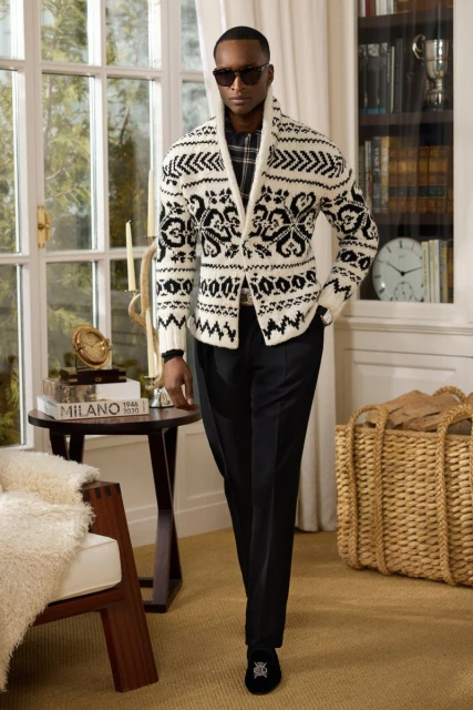 An adult Black male poses indoors wearing a patterned white sweater, dark trousers, and sunglasses, standing near a window and table.