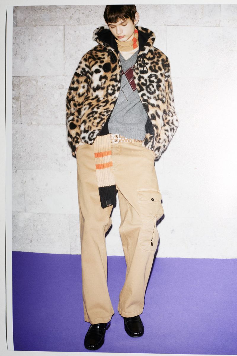 Model wearing a leopard print hooded jacket, grey patterned sweater, and beige cargo pants against a stone wall.