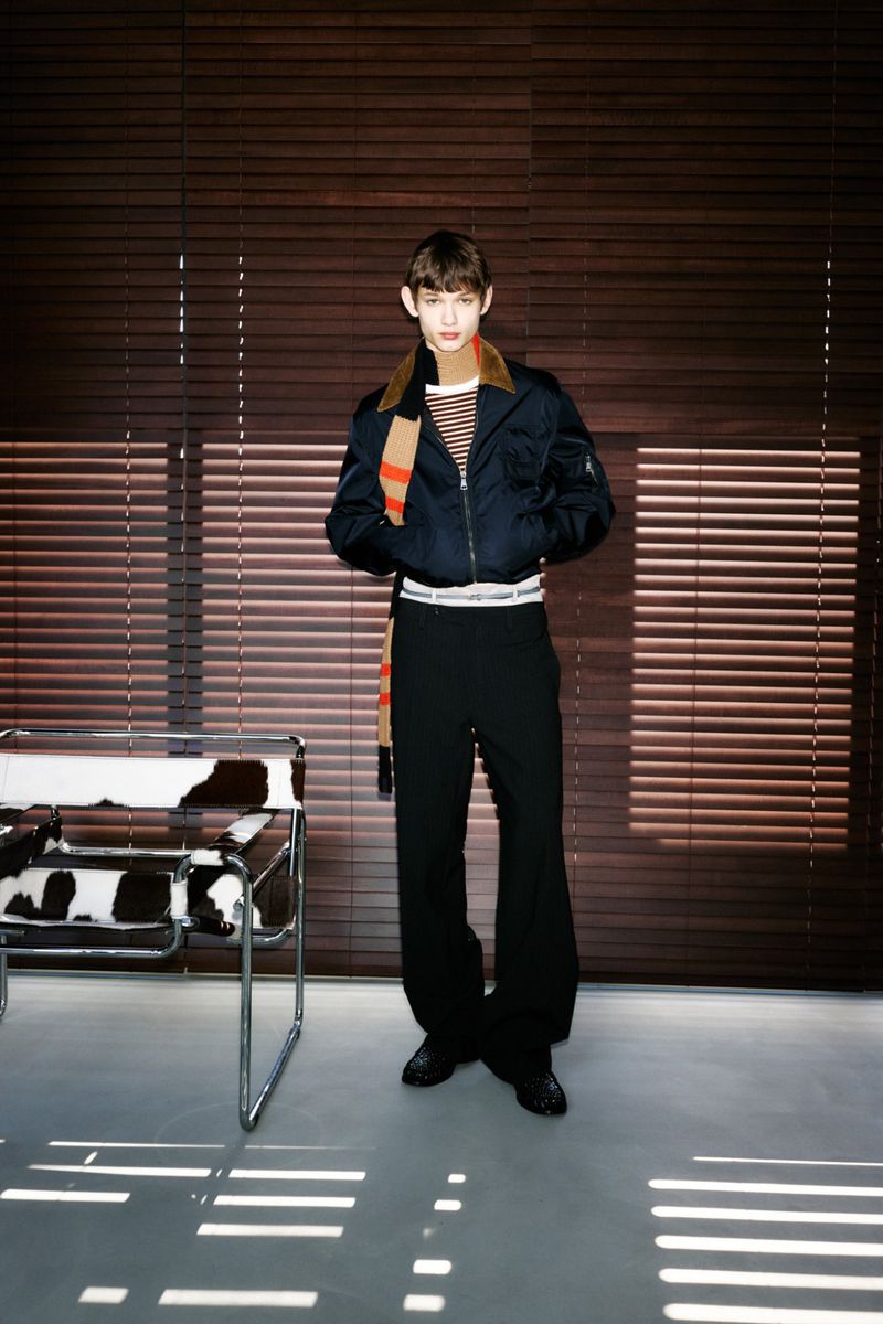 Young adult posing indoors against wooden blinds in a striped shirt, black bomber jacket, black pants, and scarf; chrome chair on the side.
