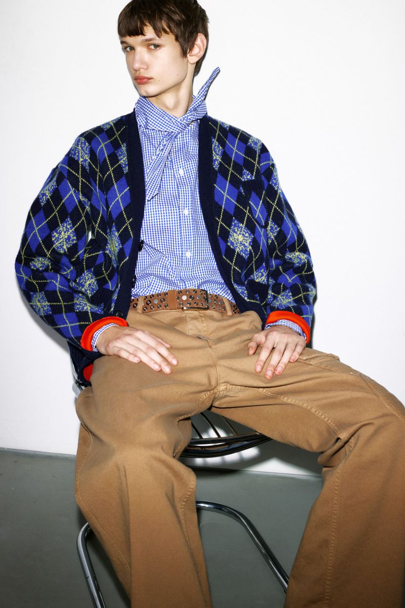 Man sits on a chrome stool in a blue patterned cardigan, light blue checked button-down shirt, tan pants, and a brown belt.