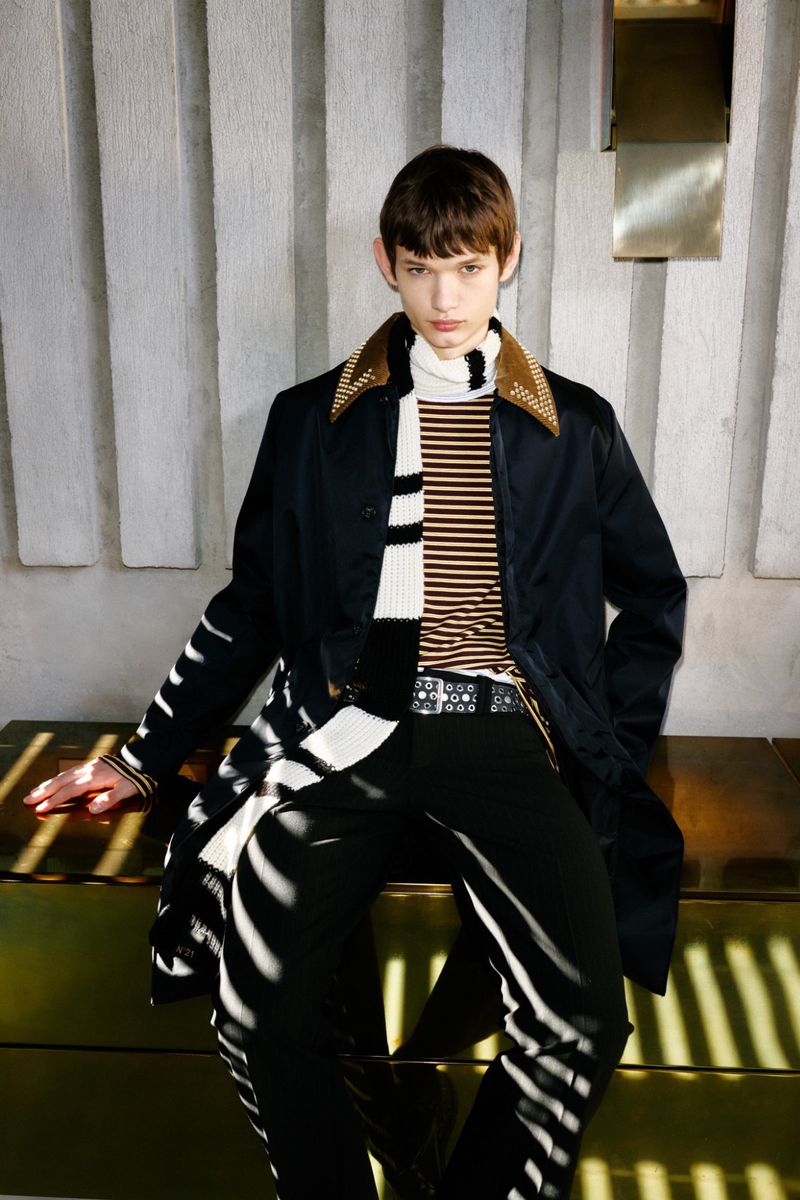 A young adult with brown hair is posing in a fashionable outfit with a black coat, a scarf, and black pants in an indoor setting.