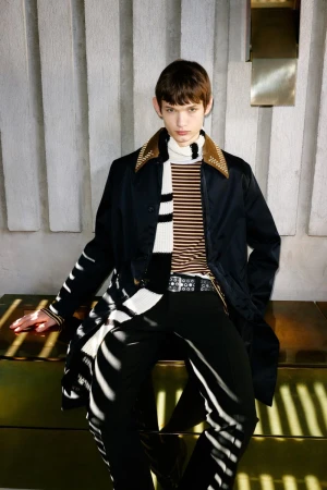 A young adult with brown hair is posing in a fashionable outfit with a black coat, a scarf, and black pants in an indoor setting.