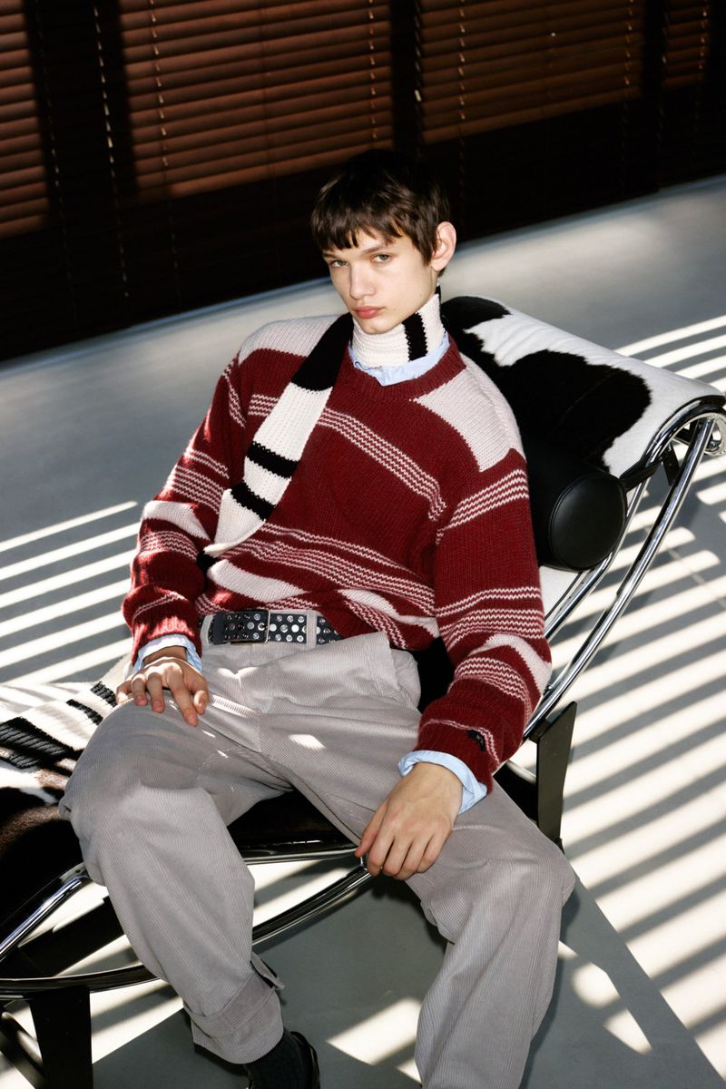Stylish young man poses while sitting in chair wearing a red and white striped sweater, grey pants, and black shoes with slatted window blinds behind.