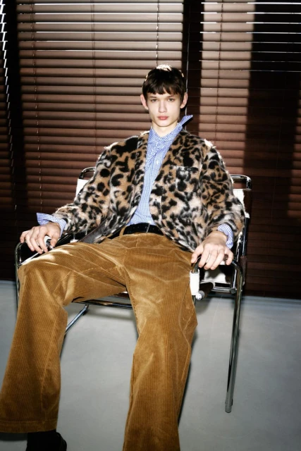 A young adult male is sitting in a metal chair in front of window blinds, wearing a patterned faux fur jacket, dress shirt, and corduroy pants.