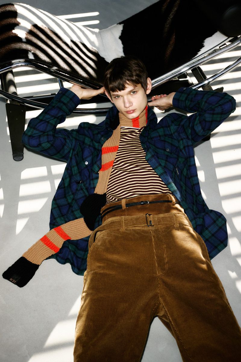 Young adult poses with a blue and green plaid shirt and brown corduroy pants on a gray surface near a metal and zebra-print chair.