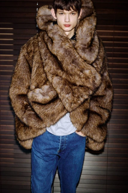 A young male model with brown hair poses in a large brown fur coat and blue jeans against a dark brown wood paneled wall for a fashion shot.