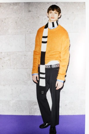 Model wearing orange crewneck sweater and striped scarf standing against a stone wall for N21 F/W 25 Men's Lookbook.